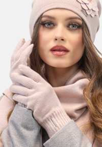 Guanti lavorati a maglia rosa chiaro e berretto abbinato con un fiore decorativo. Texture morbida, polsini a coste, abbinati a una sciarpa rosa chiaro e a un cappotto grigio.