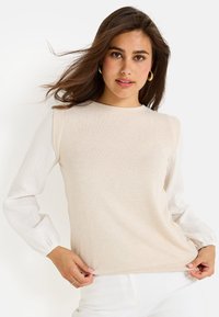 Top beige claro con un cuerpo liso y mangas largas blancas texturizadas con sutiles rayas finas. Cuello redondo y silueta ajustada.