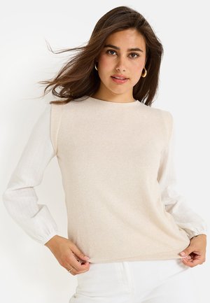 Haut beige clair avec un corps lisse et des manches longues blanches texturées présentant de subtiles rayures. Col rond et silhouette ajustée.