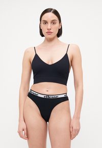 Fekete bralette vékony pántokkal és V-nyakkal, fekete, azonos színű tanga alsóval, fehér logós derékpánttal. Simább anyagtextúra.