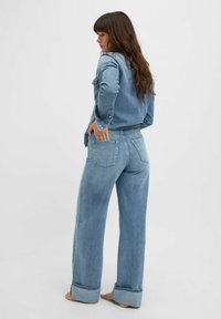 Chaqueta de mezclilla azul clara y jeans de pierna ancha, que cuentan con cinco bolsillos, un cierre frontal con botones y costuras visibles a lo largo de las costuras.