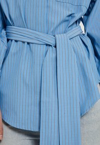 Camicia azzurro chiaro con righe bianche verticali, cintata in vita. Presenta una tasca sul lato sinistro e una texture di tessuto morbida e liscia.