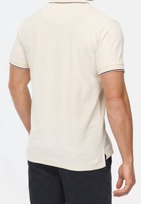 Beige polo paita lyhyillä hihoilla, jossa tummat koristeet kauluksessa ja hihoissa, valmistettu sileästä kankaasta. Saumareiät helmassa.