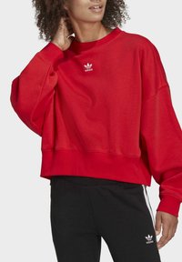 Rode cropped sweatshirt van zachte stof met geribbelde manchetten, met op de borst een witte geborduurde Adidas-logo. Gemaakt in combinatie met zwarte broek.