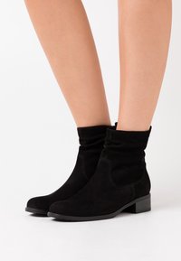 Botas de ante negras hasta el tobillo con tacones bajos, usadas sobre piernas desnudas contra un fondo blanco.