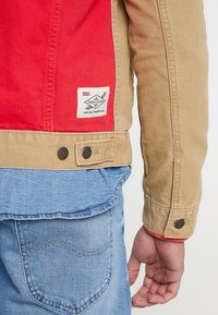 Béžová a červená denimová bunda s nášivkou Levi's, upínáním na patenty a kontrastními texturami. Viditelné švy podél lemů a rukávů.