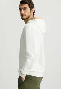 Weißer Hoodie mit einer beige gefütterten Kapuze, mit gerippten Bündchen und Saum. Glatte Textur, lässige Passform, getragen mit olivgrünen Hosen.