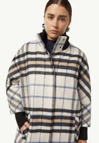 Wollmantel mit großem karierten Muster in Weiß, Schwarz und Blau. Er hat einen hohen Kragen, zwei seitliche Taschen und eine lockere Passform.