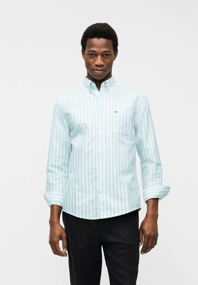 Άνδρας που φοράει πουκάμισο με κουμπιά γαλάζιο και λευκό ριγέ, με γυρισμένα μανίκια και μαύρο παντελόνι, στέκεται μπροστά σε λευκό φόντο.