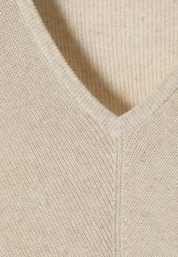 Vaalean beige värinen V-kaula-aukkoinen neulepaita. Rakenteellinen kangas, jossa näkyvät kuidut, resorikauluksella ja saumoilla, jotka muodostavat siksak-kuvion.