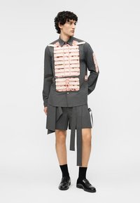 Meesmodell, kes kannab tumedat halli särki roosade lillemustritega, hallid lühikesed püksid pikkade kangast ribadega, mustad sokid ja mustad kingad.