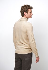 Beige polotröja med mjuk textur, ribbade ärmslut och fåll, tillsammans med mörkbruna byxor, visad bakifrån.