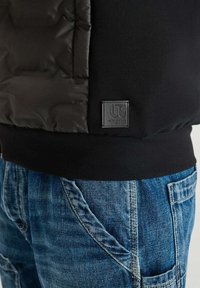 Giacca nera imbottita con una finitura liscia e testurizzata, dotata di una patch con logo e polsini a coste. Abbinata a jeans in denim blu.