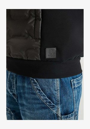 Giacca nera imbottita con una finitura liscia e testurizzata, dotata di una patch con logo e polsini a coste. Abbinata a jeans in denim blu.