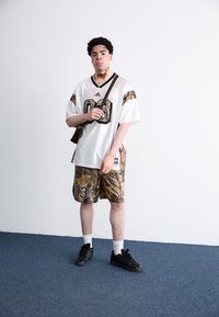 Camisa branca com detalhes em preto e dourado e o "03" na frente, combinada com calções de camuflagem castanhos. Ténis pretos e bolsa de ombro.
