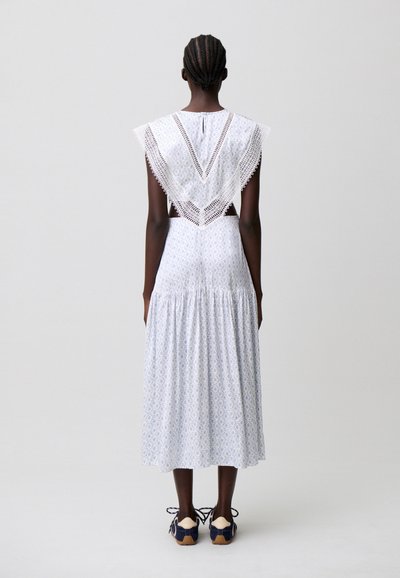 Donna con capelli intrecciati, vista di spalle, indossa un vestito midi bianco con fantasia, un mantellina bordata in pizzo e sneakers blu navy.