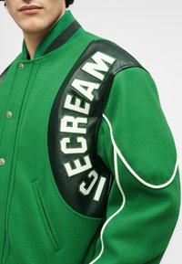 Πράσινο μάλλινο μπουφάν τύπου bomber με δερμάτινες λεπτομέρειες. Διαθέτει έντονα λευκά γράμματα, ριγέ γιακά, κουμπιά με κλίπ και πλαϊνές τσέπες.