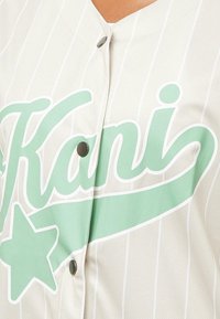 Svetlo šedý baseballový dres s vertikálnymi bielymi pruhmi a zelenou grafickou potlačou s nápisom "Kani" a hviezdou. Obsahuje čierne zapínanie na patentky.
