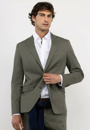 Hombre ajustando un botón en una chaqueta de traje verde oliva llevada sobre una camisa blanca, sobre fondo liso.