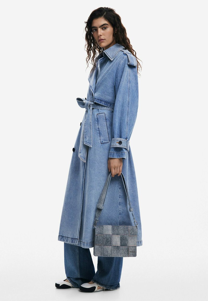 Trench coat in denim chiaro con cintura, grande colletto e tasche, abbinato a jeans a gamba larga e una borsa a toppe grigia.