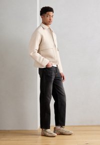 Beige ullblanding jakke med frontlommer, kombinert med mørke jeans og lyse sneakers. Strukturert materiale og enkel design.