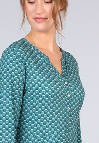 Türkis gemusterte Bluse mit V-Ausschnitt, langen Ärmeln und Knopfdetails an der Vorderseite. Mit einem Blattmotiv in verschiedenen Farbtönen.