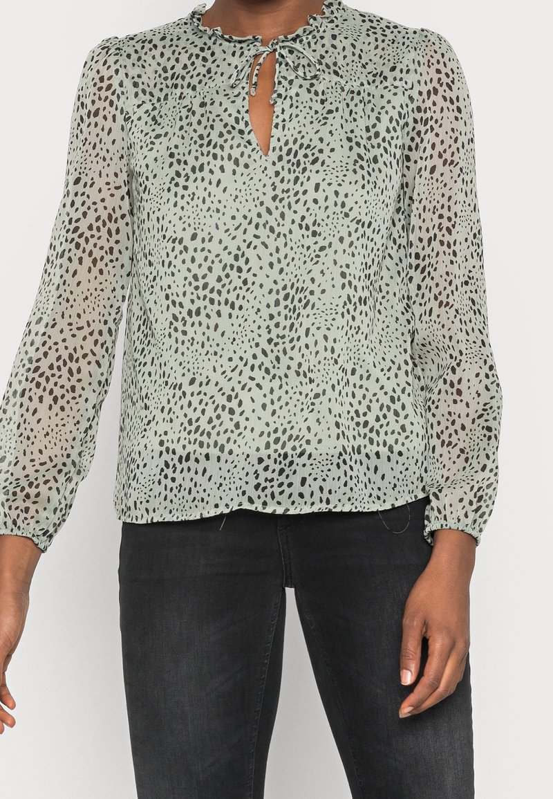 Femme portant un chemisier vert clair à manches longues avec des taches noires irrégulières et un détail noué au col, associé à un jean noir.