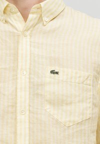 Chemise pour homme à rayures verticales jaunes et blanches, boutonnée sur le devant avec une poche ornée d'un petit logo de crocodile vert.
