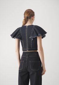 Svart denim topp med korte ermer og volanger, med glidelås bak og kontrasterende sømmer. Brukt sammen med høytlivs svarte bukser.