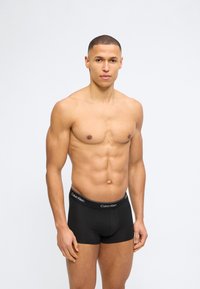 Μυώδης άνδρας με κοντά μαλλιά, φορώντας μαύρα εσώρουχα Calvin Klein τύπου boxer, στέκεται μπροστά σε απλό λευκό φόντο, κοιτάζοντας μπροστά.