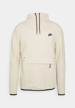 Krémově zbarvený Nike pulovr s vysokým límcem, černými stahovacími šňůrkami, přední kapsou na zip a černým logem na levé straně hrudi.