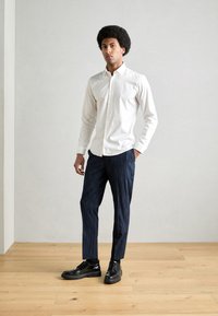 Hvit skjorte med knapper, slim-fit bukser med marineblå stipestriper og sorte lærersko. Glatt stoff, skreddersydd design og definerte linjer.