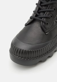 Gros plan sur une botte noire avec une semelle en caoutchouc texturée, un bout rond et un devant à lacets avec œillets métalliques.