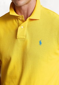 Gul polotröja i strukturerad tyg med en tvåknäppningsknäppning; har en blå broderad logotyp på vänster bröst.