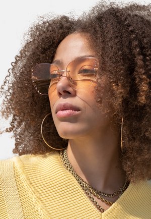 Rose-tinted, oversized sunglasses with metal accents and clear frames; worn with a yellow textured sweater and layered gold necklaces.