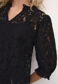 Black lace blouse with floral pattern, v-neckline, and puffed sleeves. Delicate texture with scalloped edges at collar and cuffs.