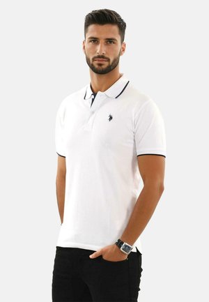 Weißes Poloshirt mit marineblauem Besatz am Kragen und an den Ärmeln, mit einem kleinen Logo auf der Brust. Getragen mit schwarzen Hosen.
