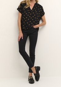 Mujer vistiendo una blusa negra de manga corta con lunares marrones, jeans ajustados negros y mocasines gruesos negros con detalle dorado.