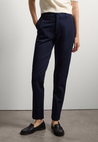 Marineblauwe, op maat gemaakte broeken van een gladde stof, met zijzakken en een slim fit ontwerp. Gecombineerd met zwarte platte instappers.