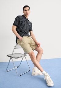 Kortærmet sort polo med geometrisk mønster, beige shorts og hvide sneakers med et logo. Sidder på en krom foldebænk mod en blå baggrund.