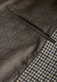 Pruunist mustrist kanga on geomeetriline muster, millele on lisatud tekstuuriga houndstooth muster beeži ja tumeläbi värviga.