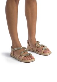 Sandali piatti beige con cinturini intrecciati, dotati di accenti metallici e strass decorativi. Sole testurizzato con una leggera lucentezza.