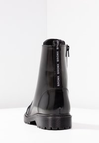 Botín negro brillante hasta el tobillo con suela gruesa y contundente, cremallera trasera y tirador etiquetado con "BRONX" en texto blanco, fotografiado sobre fondo blanco.