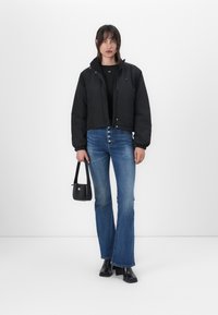 Svart cropped jakke med høy krage, kombinert med blå utvidede denimjeans med knappestolpe. Tilbehør inkluderer en liten svart håndveske.