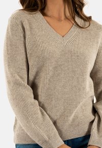 Donna in maglione beige a coste con scollatura a V e jeans blu, che mostra la parte superiore del torso e una parte delle braccia contro uno sfondo bianco.