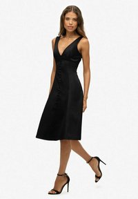 Schwarzes A-Linien-Kleid mit V-Ausschnitt, mit Knopfleiste an der Vorderseite, ärmellosem Design und glattem Stoff, kombiniert mit schwarzen High-Heels.