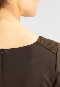 Ciemnobrązowy top z żebrowanym akcentem na ramieniu oraz małym zamkiem z tyłu. Gładka tekstura materiału w strukturalnym fasonie.