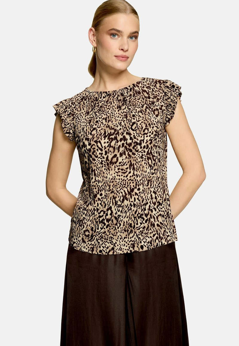 Femme aux cheveux blonds portant un chemisier sans manches à motif léopard avec des épaules à volants et un pantalon ample foncé sur un fond uni.