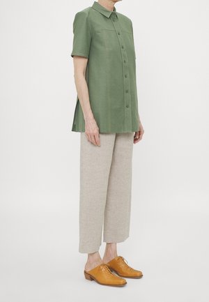 Camicia a maniche corte verde con bottoni e taglio dritto, abbinata a pantaloni in lino beige chiaro e scarpe stringate in pelle marrone.