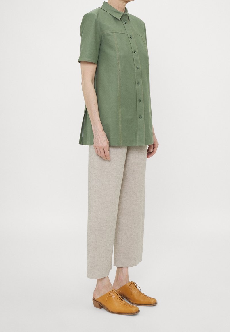 Camicia a maniche corte verde con bottoni e taglio dritto, abbinata a pantaloni in lino beige chiaro e scarpe stringate in pelle marrone.